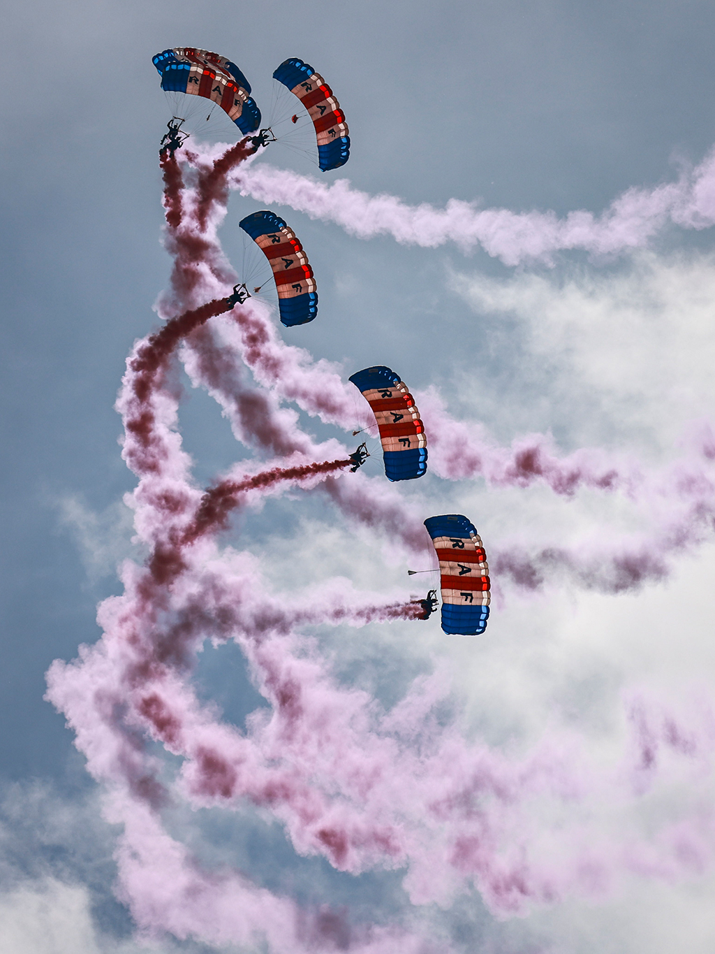 Six RAF Falcons Parachute Display Team members descend in a vertical formation, beneath their distinctive red, white, and blue canopies, which each spell out "RAF" on their white panels, while leaving spiralling trails of red smoke in a cloudy sky.